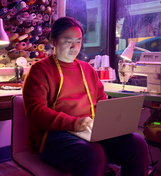 Eine Person nutzt ein MacBook Neo in Rosa auf dem Schoß, das nicht am Strom angeschlossen ist, in einem Arbeitsbereich mit einer Nähmaschine und Zubehör wie Garn und einer Schneiderpuppe